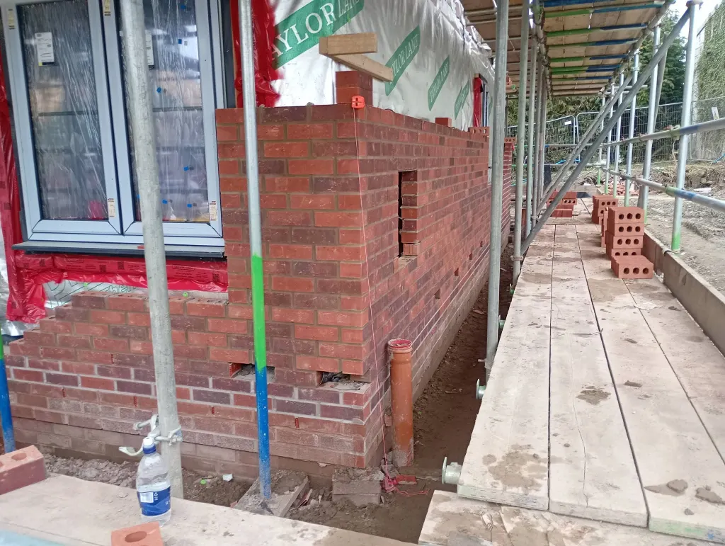 Corner of red facing brick racked back beside a window, with blue lower courses and a long scaffold walkway behind