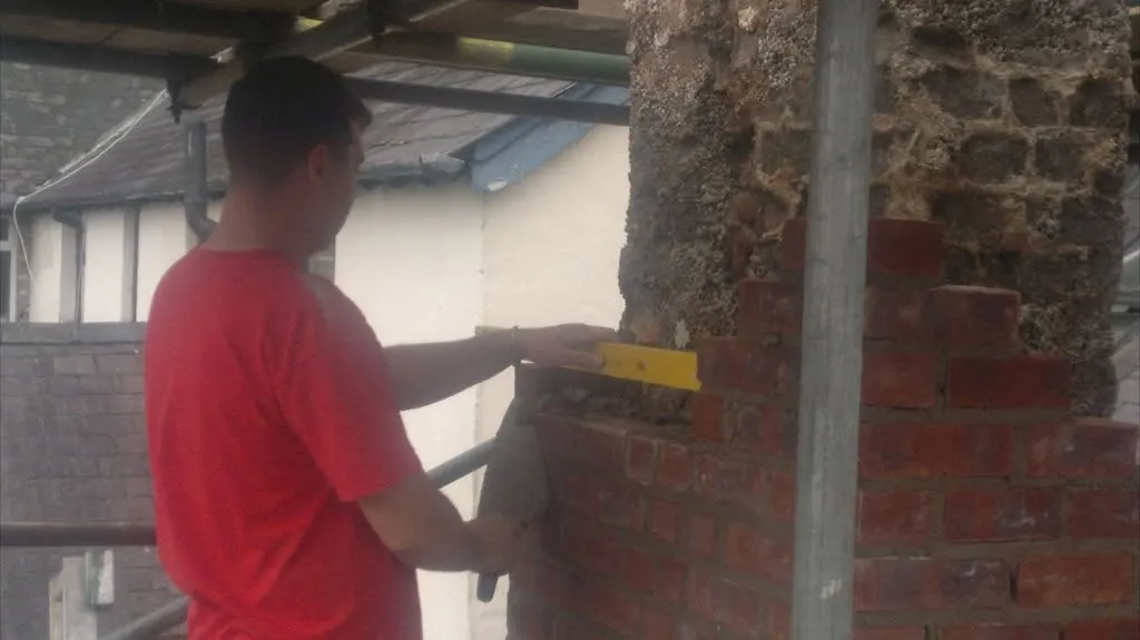 Bricklayer in a red T-shirt holding a spirit level to new brickwork built against an old stone chimney on a scaffold