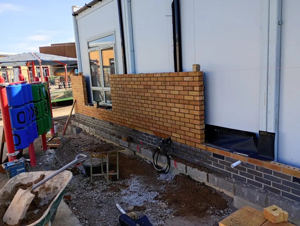 Buff facing brick panel being built under a window against white wall panels, with play equipment in the yard beyond