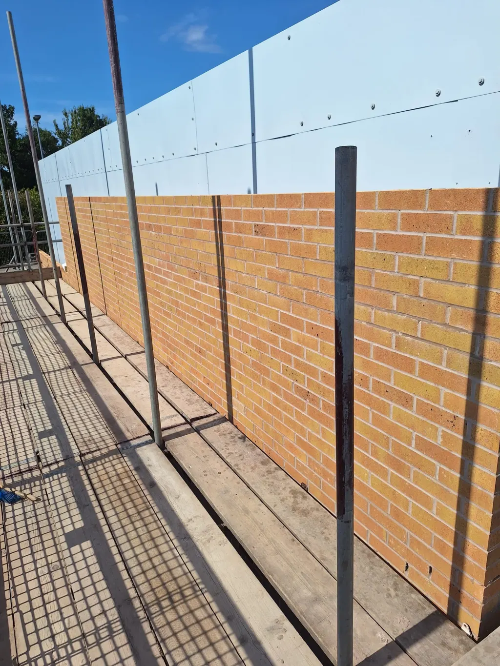 Buff facing brick wall built up beneath white wall panels, seen from the scaffold with standards along the edge