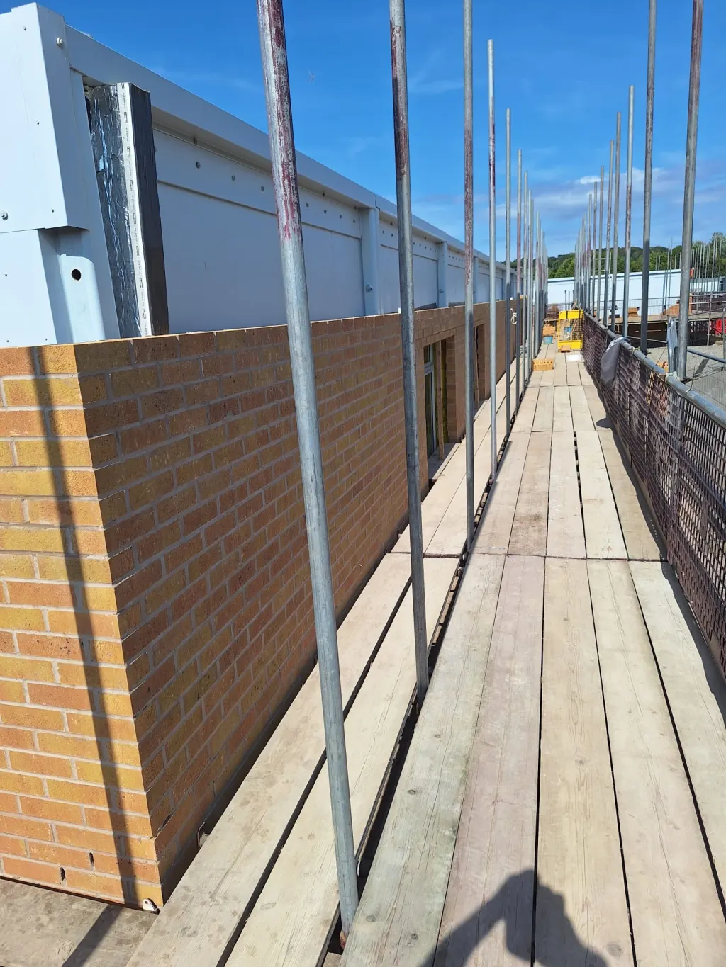 Buff facing brick wall built against a white-clad building, seen along clean scaffold boards on a sunny day