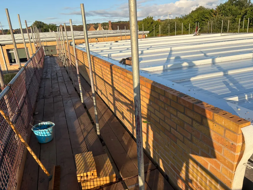 Buff facing brick wall built off scaffold beside a pale metal roof, with bricks, a tub and a bucket on the boards