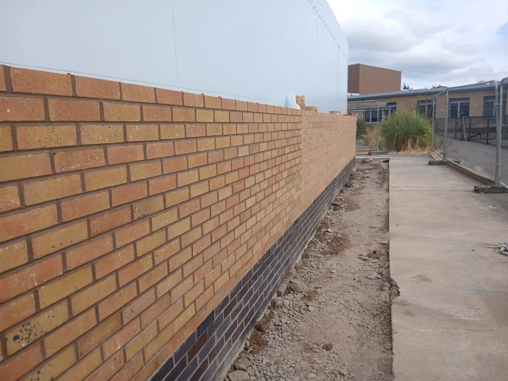 Buff facing brick wall on a dark engineering brick base course, built up against smooth white wall panels