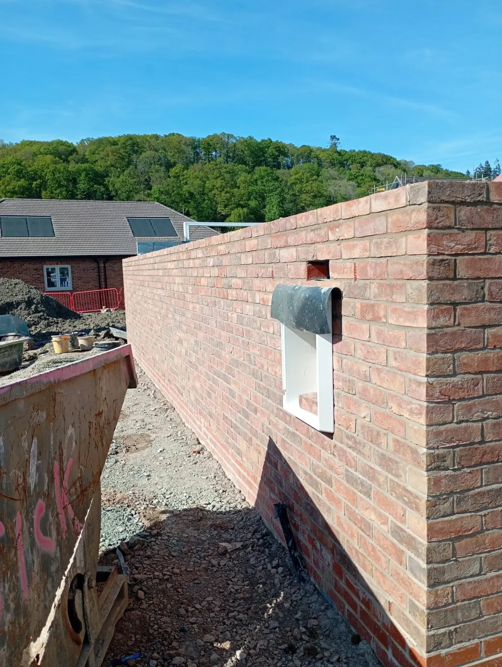 Plain red facing brick wall with a meter box recess and flashing, a site skip in front and a wooded hillside behind