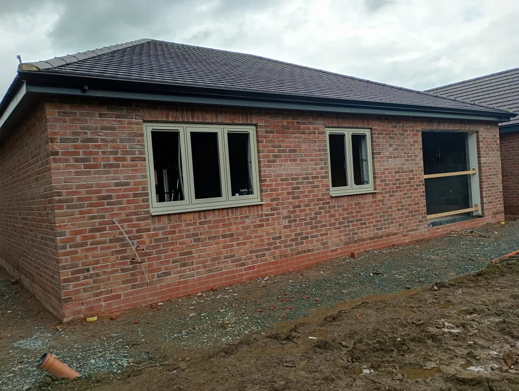 Rear of a new bungalow in red multi facing brick with cream windows, a hipped tiled roof and bare ground in front
