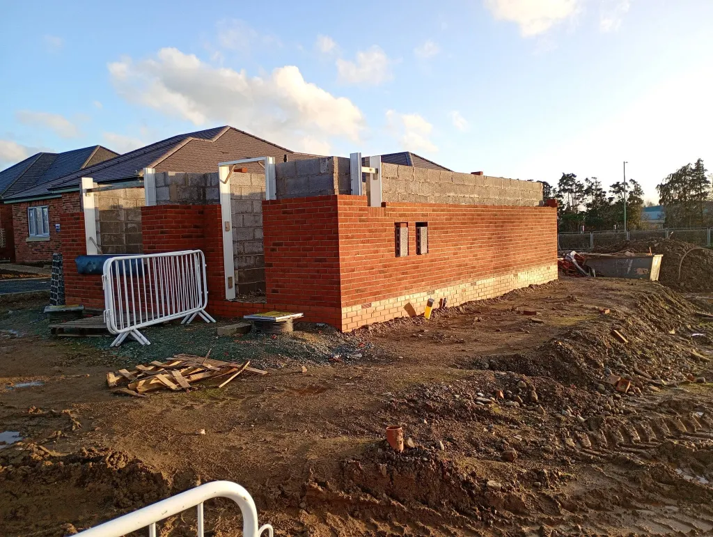 Bungalow shell in red facing brick at window-head height, with blockwork above and completed bungalows behind