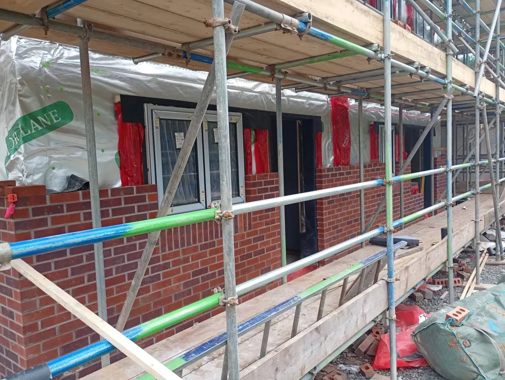 Red facing brick built up to window sill height beneath a fitted window, seen through scaffold tubes and boards