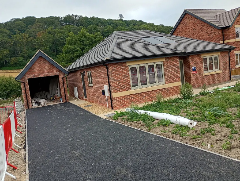 Finished bungalow in red brick with a buff stone band, a detached garage and a new tarmac drive, wooded hills behind