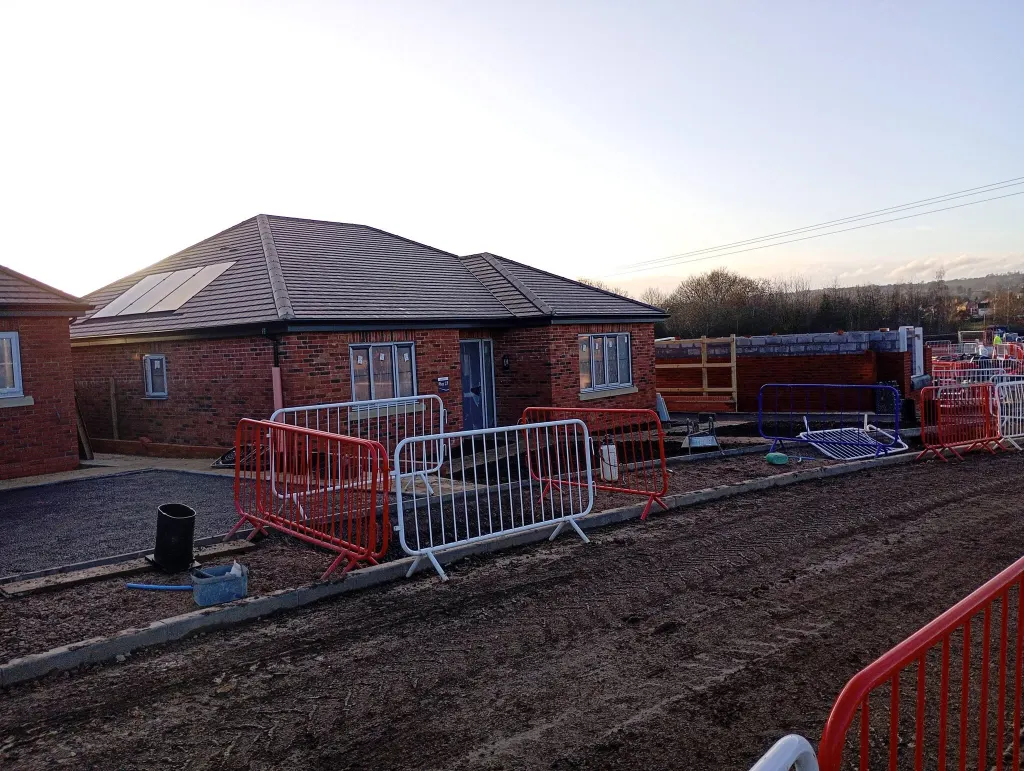 Pair of completed brick bungalows with hipped roofs in low evening light, behind red and white site barriers