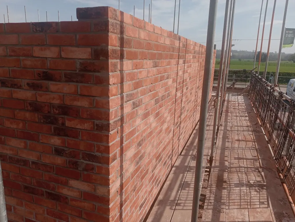 Tall corner of multi red facing brick built plumb and to gauge, with a scaffold walkway and fields alongside