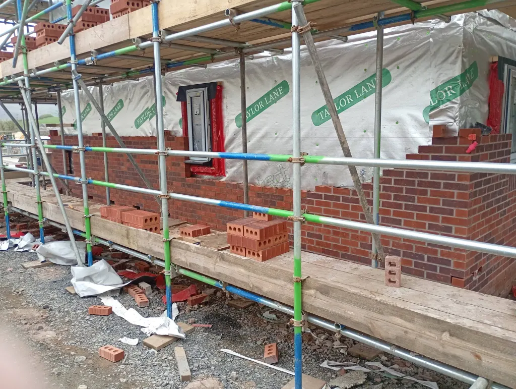 Facing brick outer leaf rising off scaffold below a window, with membrane-wrapped frame above and bricks loaded on boards