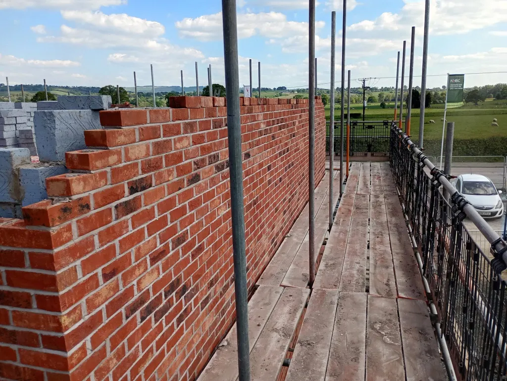 Facing brick wall racked back along a scaffold walkway, with blockwork behind and green fields in the background
