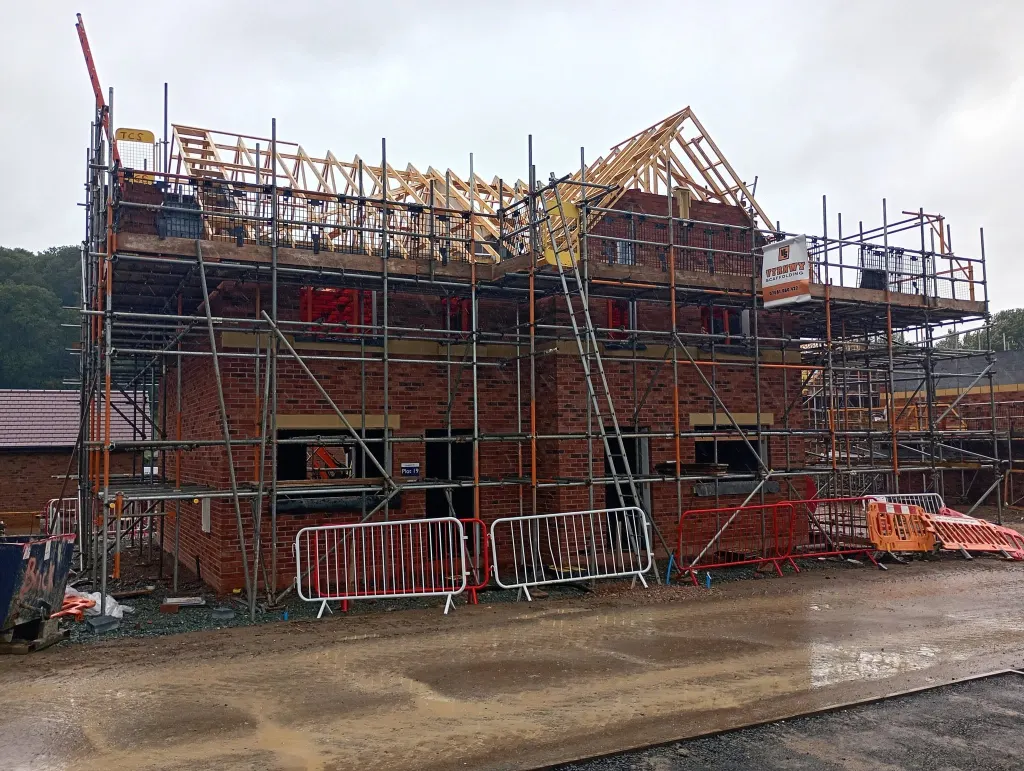 Brick house under full scaffold with timber roof trusses going on, site barriers and a wet road in front