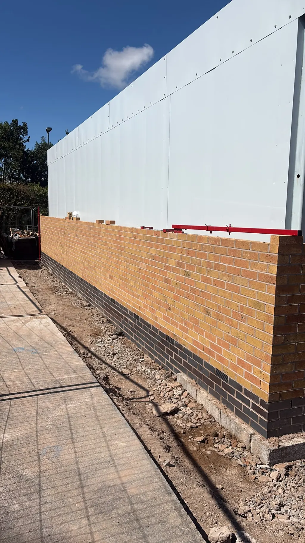 Long run of buff facing brick with a dark brick base, built up beneath white wall panels beside a block-paved path