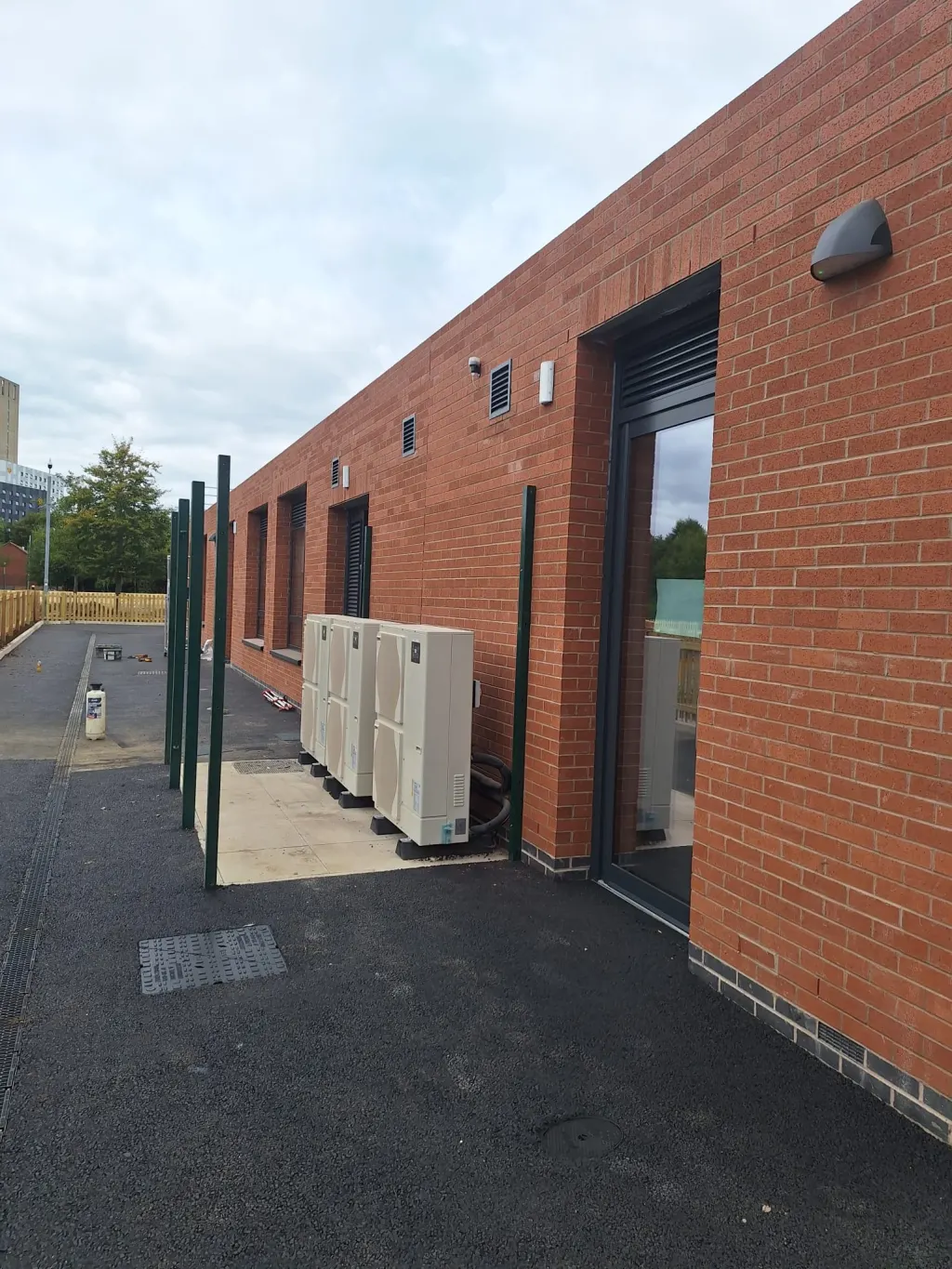 Single-storey building in red facing brick with a glazed door and wall light, heat pump units along the tarmac path