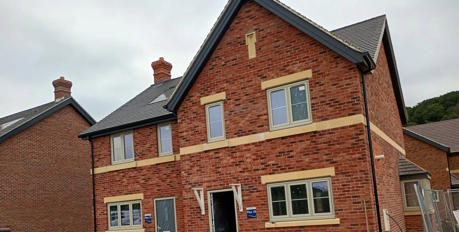 Semi-detached houses in red brick with a front gable, buff stone bands, chimneys and grey tiled roofs on a new estate