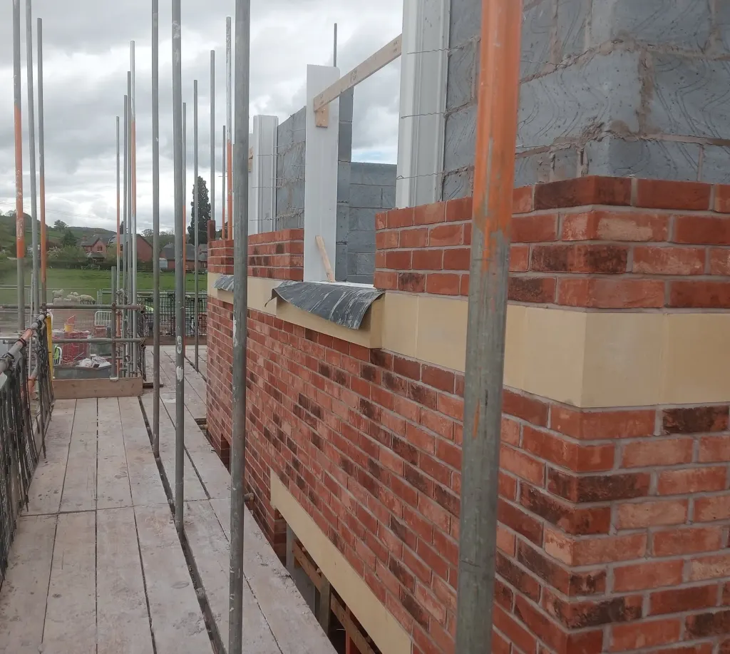Buff stone band and sills set in red facing brick at first floor, window frames propped and sheep in the field beyond