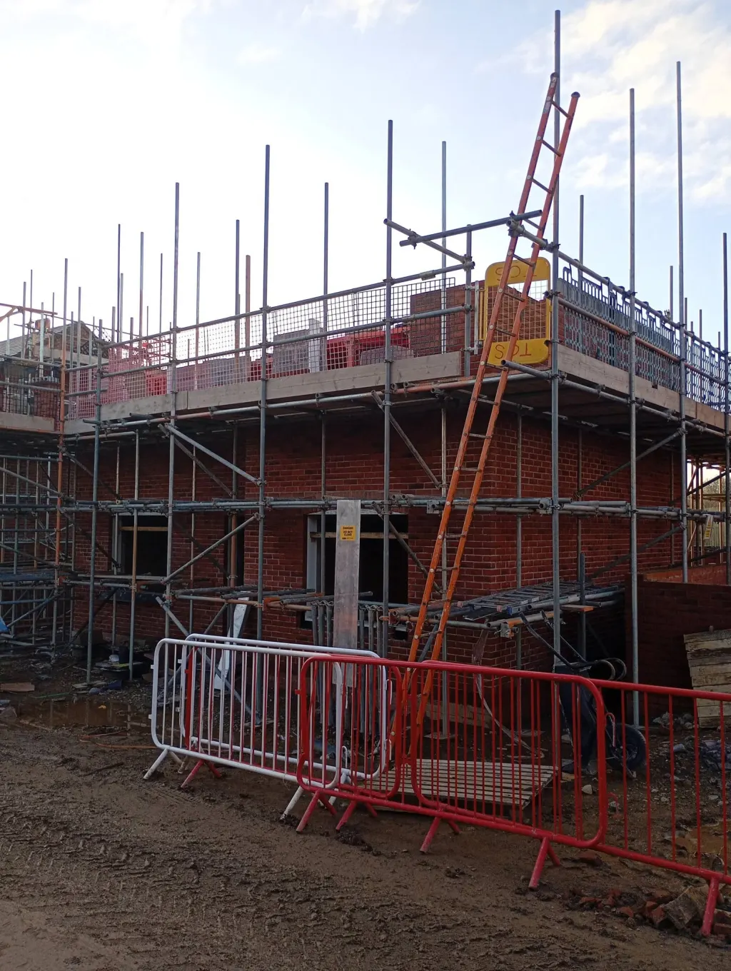 Two-storey house in red facing brick wrapped in scaffold, with an orange ladder, loading gate and site barriers in front