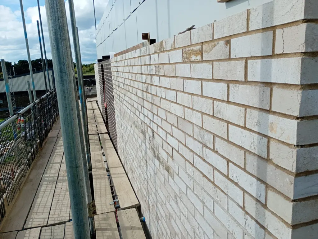 Corner of white-grey facing brick built off the scaffold, with the walkway, tubes and handrail running alongside